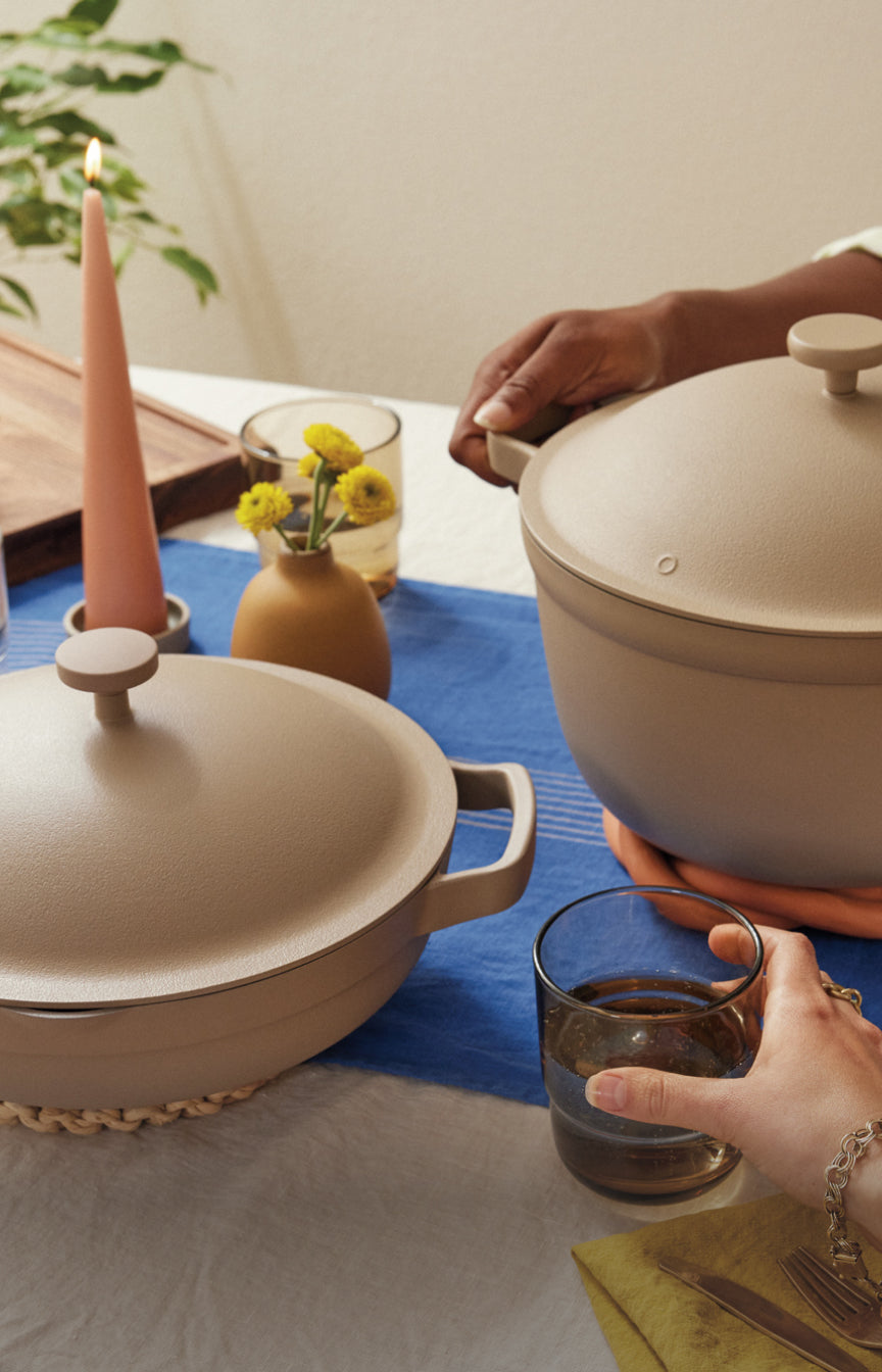 Two hands are shown at a dining table, one lifting the lid off a beige pot, the other holding a glass of water. On the table are another covered pot, a pink candle, yellow flowers in a vase, and a blue table runner.