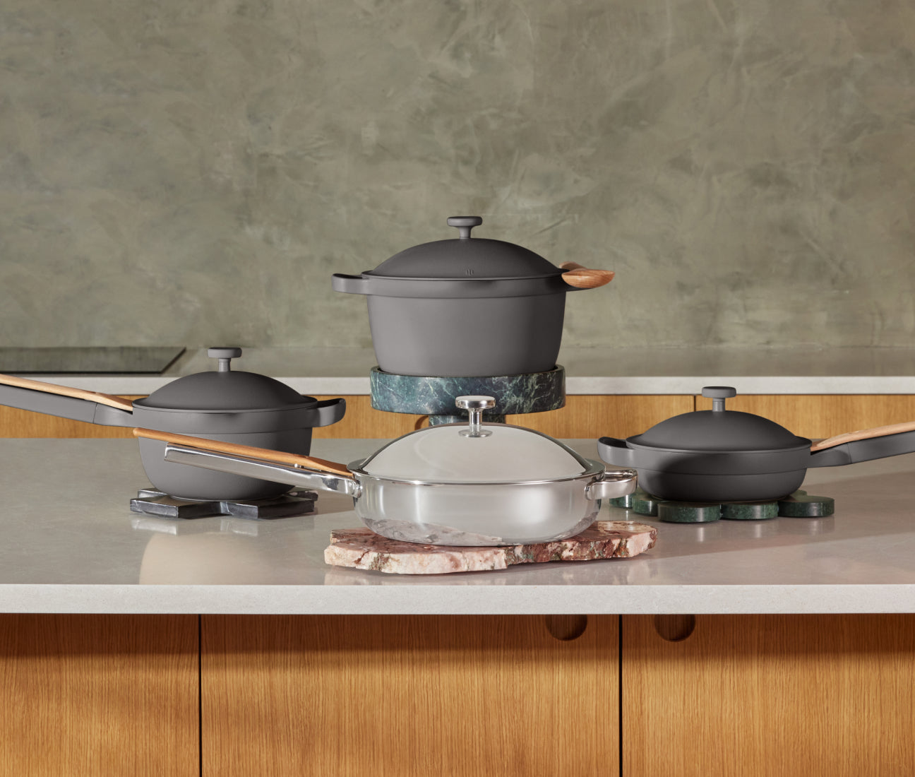 A set of modern cookware including two gray pots, a gray pan, and a silver pan with wooden handles, arranged on a kitchen counter with a greenish textured wall in the background.