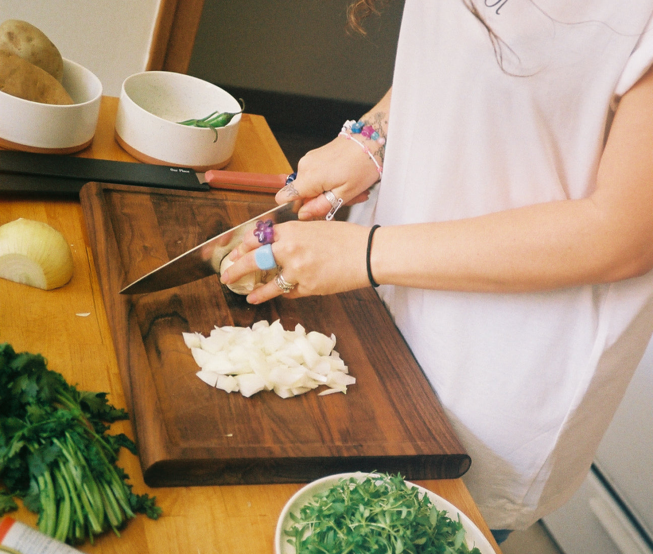 A person wearing rings and bracelets is chopping an onion on a wooden cutting board. Fresh herbs and vegetables are visible on the table, along with bowls and a knife.