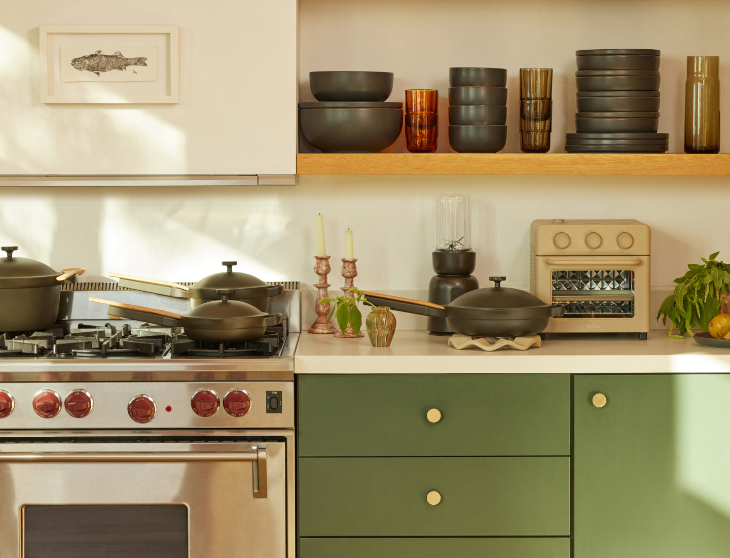 A modern kitchen with green cabinets, a stainless steel oven, cookware on the stove, and shelves displaying stacked dishes, glasses, and a small toaster. A framed fish drawing hangs on the wall. Sunlight brightens the scene.