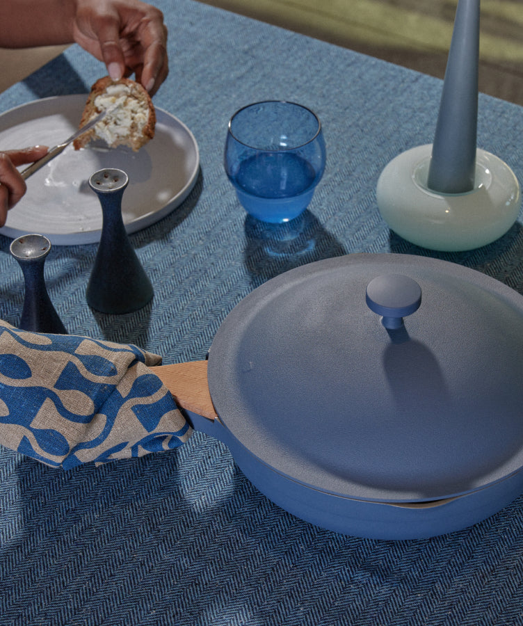 A table set with blue dishes, a covered pot, a glass of water, a bowl of grapes, salt and pepper shakers, and a hand holding a wooden spatula wrapped in a patterned cloth on a blue textured tablecloth.