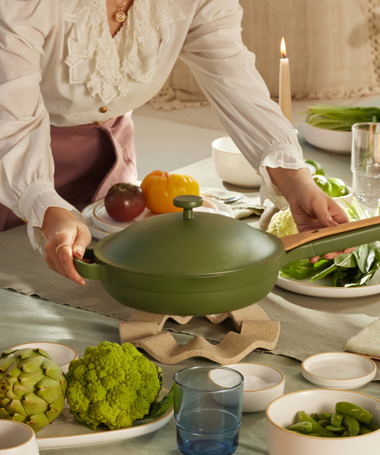 A person places a green, lidded pan with a wooden handle on a trivet at a dining table set with plates, vegetables, cups, and candles in warm natural light.