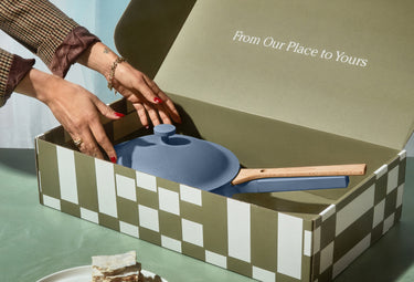A person places a blue pan with a wooden handle into a patterned gift box that reads From Our Place to Yours. The box sits on a green surface next to a plate with a piece of cake.