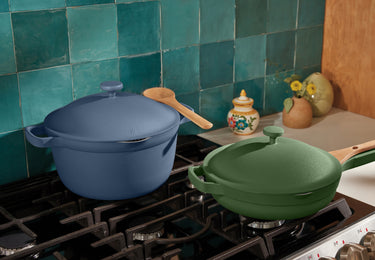 Two colorful pots, one blue and one green, with wooden handles and lids sit on a gas stove. The background features teal tiled walls and a countertop with a small vase, a ceramic jar, and yellow flowers.