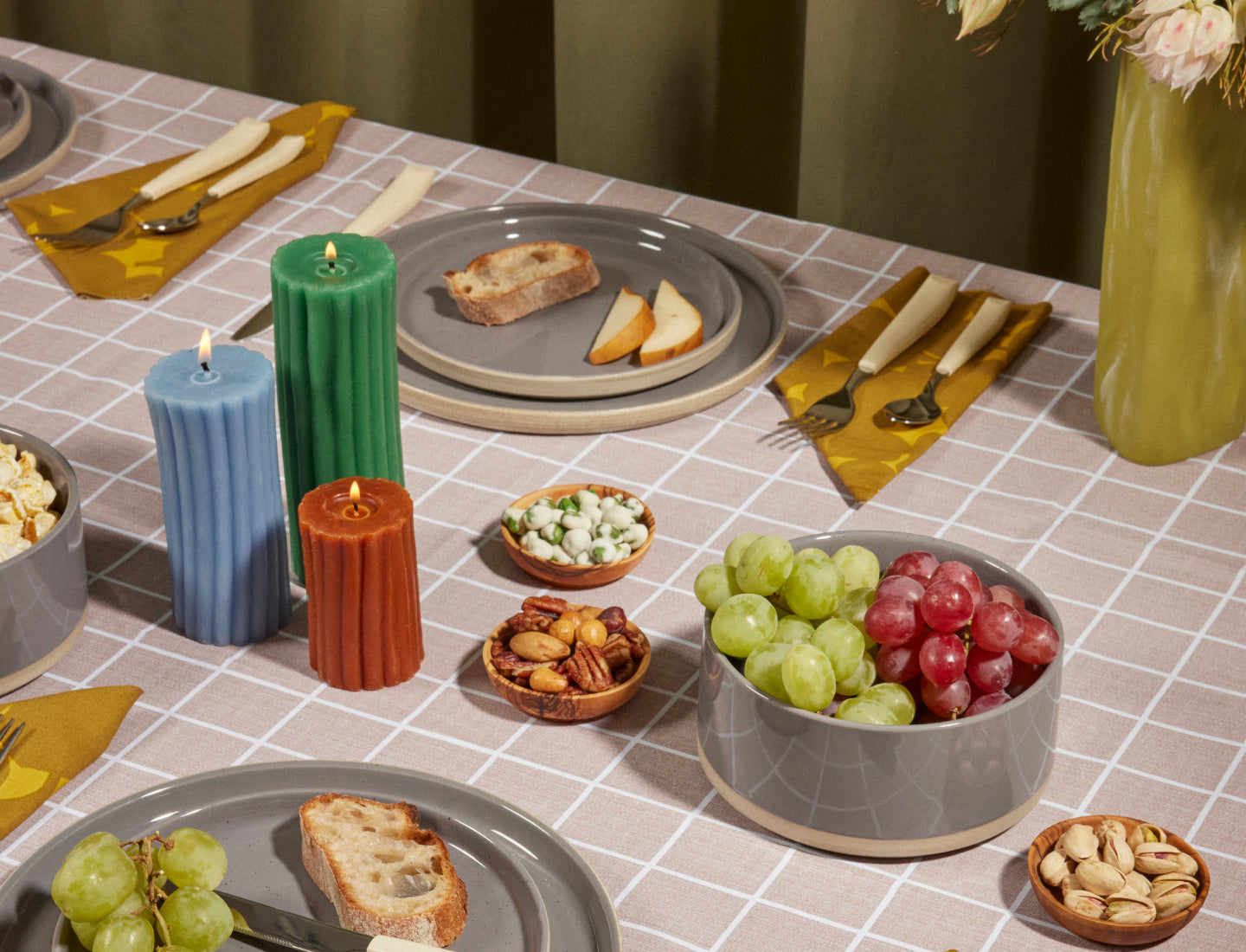 A neatly set dining table with gray plates holding slices of bread and pear, yellow napkins, assorted grapes, nuts, and snacks in bowls, three candles in blue, green, and orange, and a vase with light flowers.