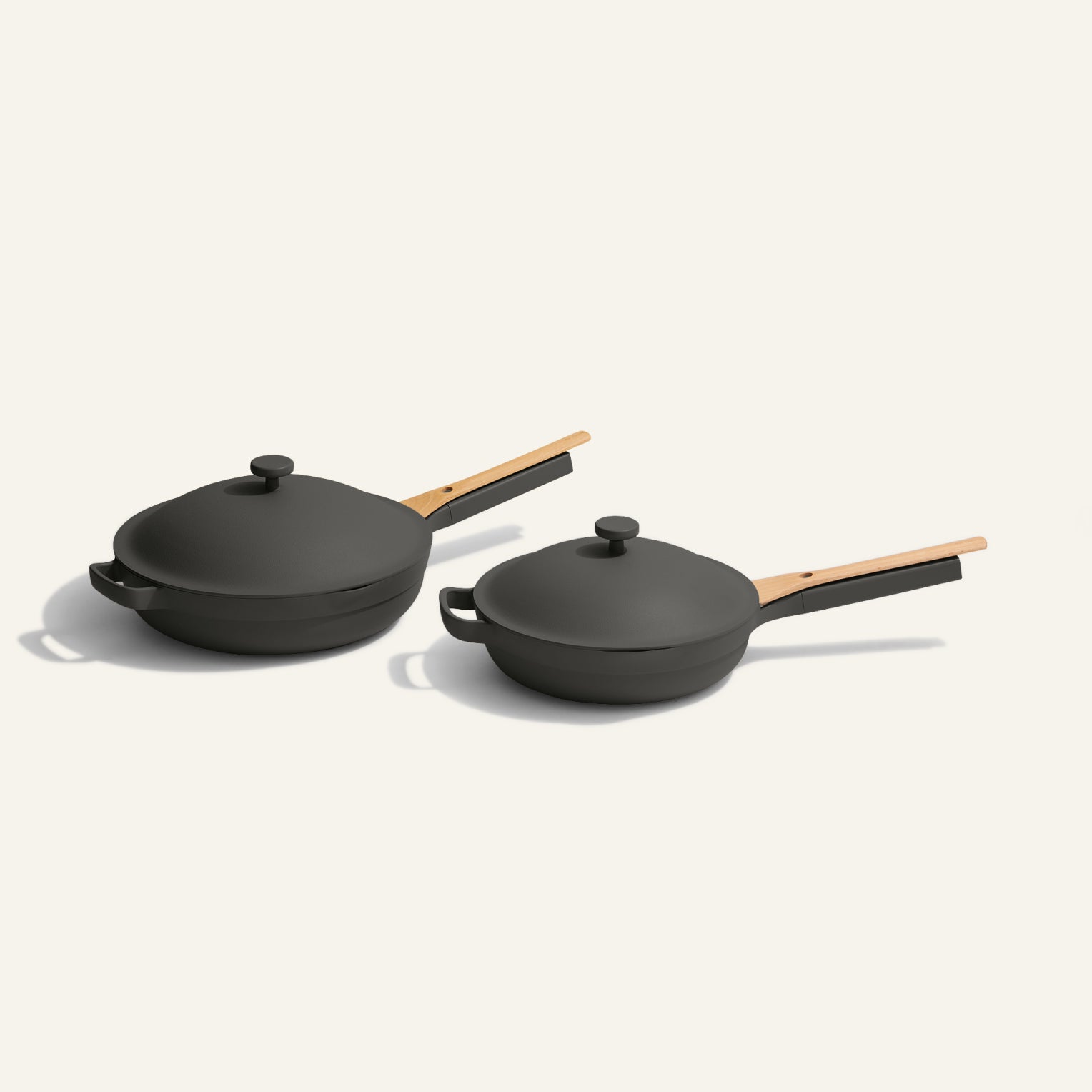 Two black nonstick pans with wooden handles and matching lids are placed side by side on a light, neutral background. One pan is slightly larger than the other.