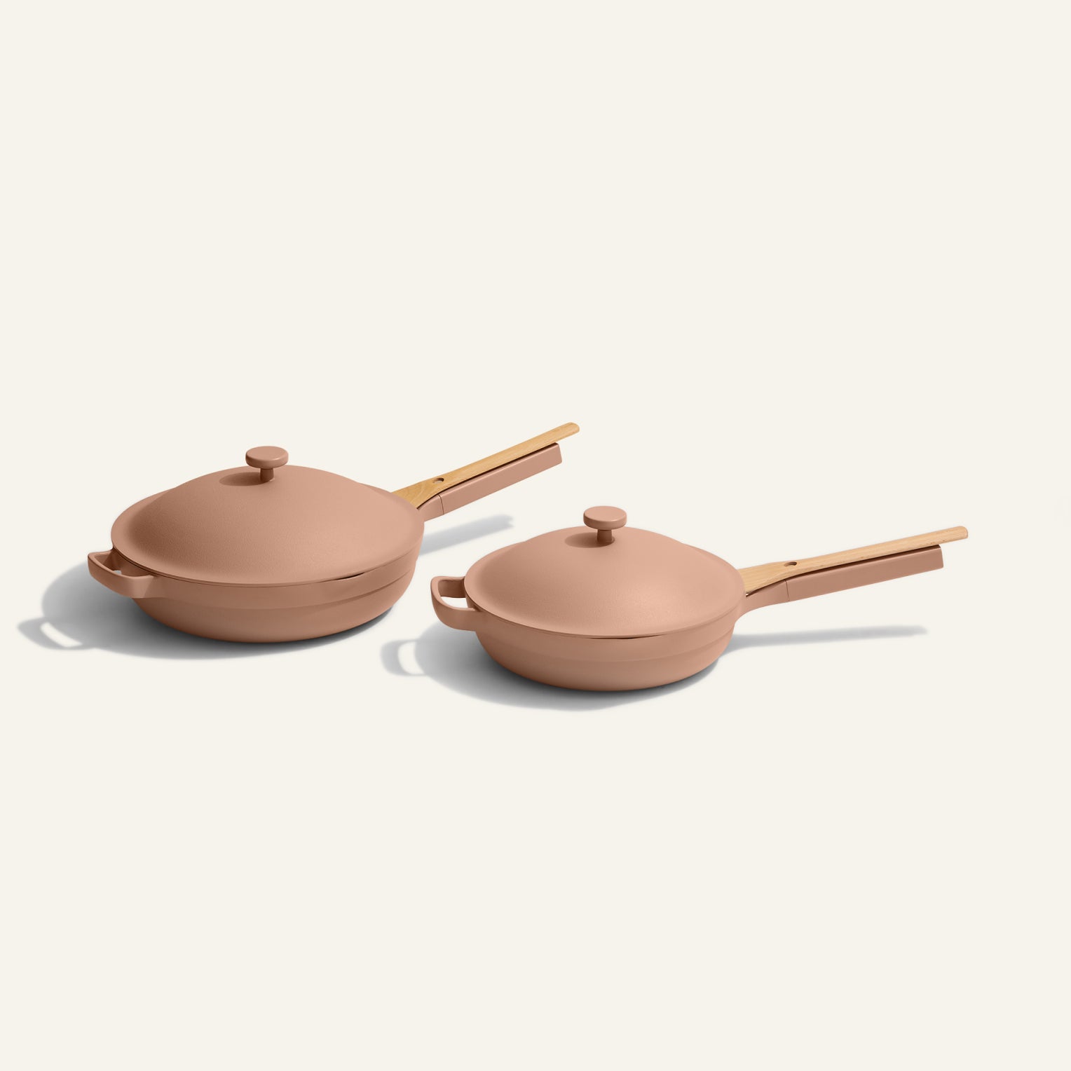 Two nonstick frying pans with matching lids and wooden handles, both in a muted pink color, are placed side by side on a light background.